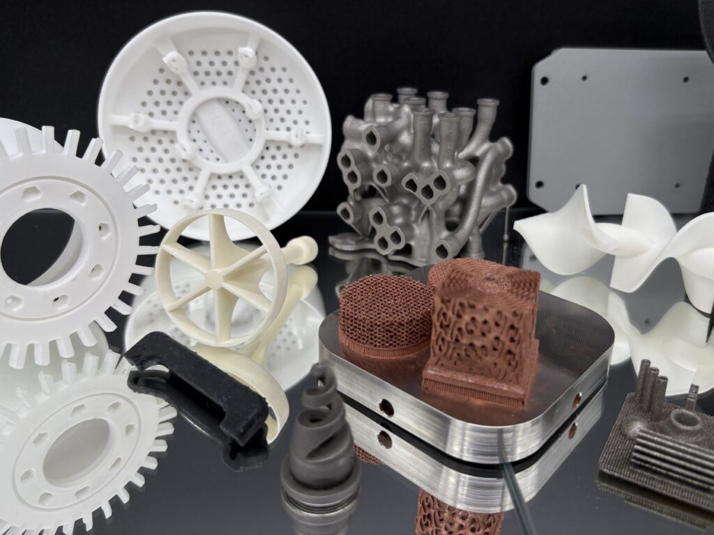 3D‑printed metal parts made from strong industrial materials, including intricate lattice structures and complex geometric components, displayed on a reflective surface.