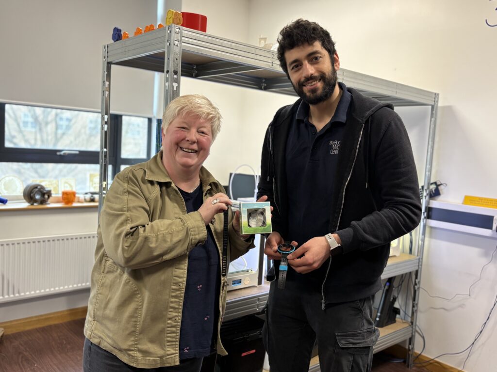 Two people standing smiling in a workshop space holding a small watch strap packaged up in a box to demonstrate how Myndtime began at the 3M BBIC.