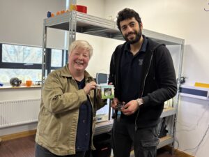Two people standing smiling in a workshop space holding a small watch strap packaged up in a box to demonstrate how Myndtime began at the 3M BBIC.